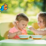 Two little girls sitting at a table and eating together against green lawn