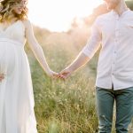 Young happy romantic pregnant couple walking in wild field in summer day. Pregnant woman in white dress and her handsome man expecting a baby, enjoying the walk in summer field