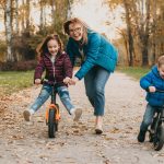 Smiling caucasian mother is playing with her small kids while teaching them to ride the bike